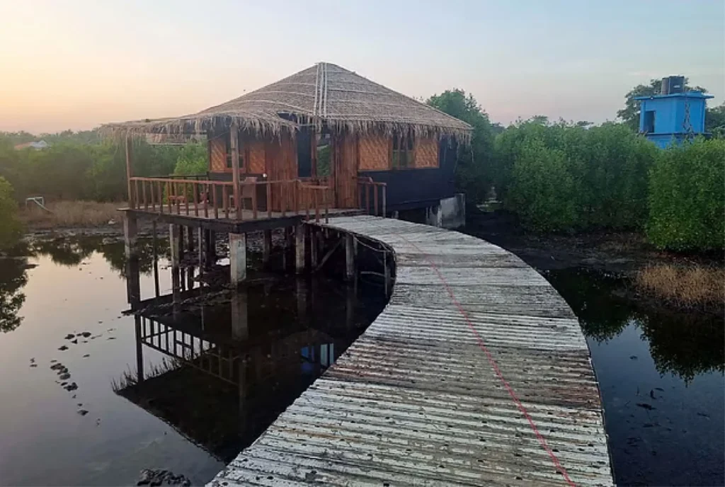 জলরাশির উপরে একটি ছনের ছাউনি দেওয়া ইকো-রিসোর্টের কুঁড়েঘর, যার দিকে কাঠের সেতু চলে গেছে।