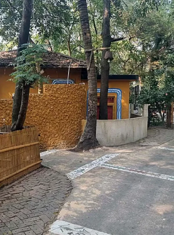 গাছপালা ঘেরা একটি মাটির বাড়ির ছবি, যার সামনে বাঁশের বেড়া এবং একটি হাঁটার রাস্তা দেখা যাচ্ছে।